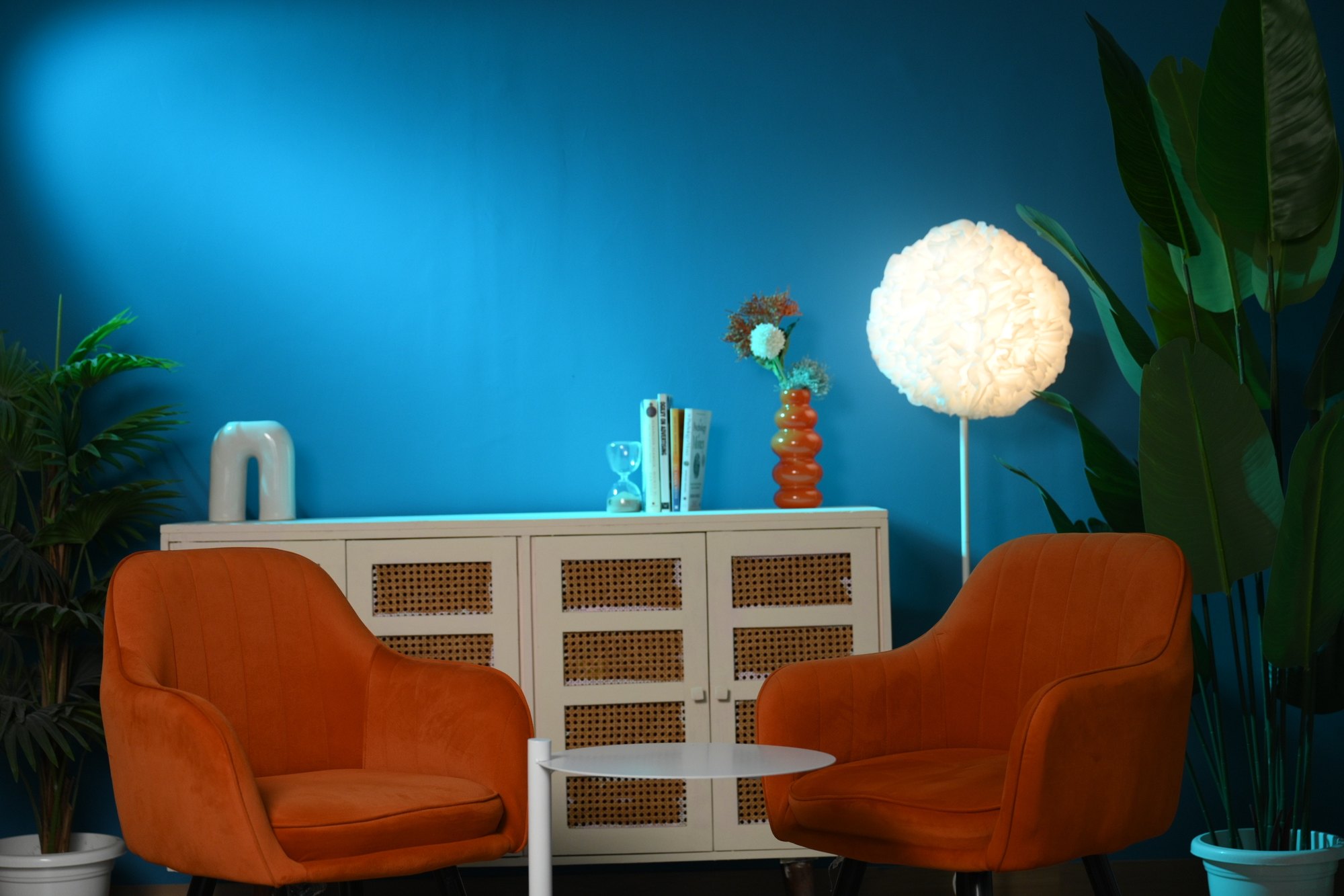 Styled teal studio wall with orange chairs plants cabinet and practical lamp